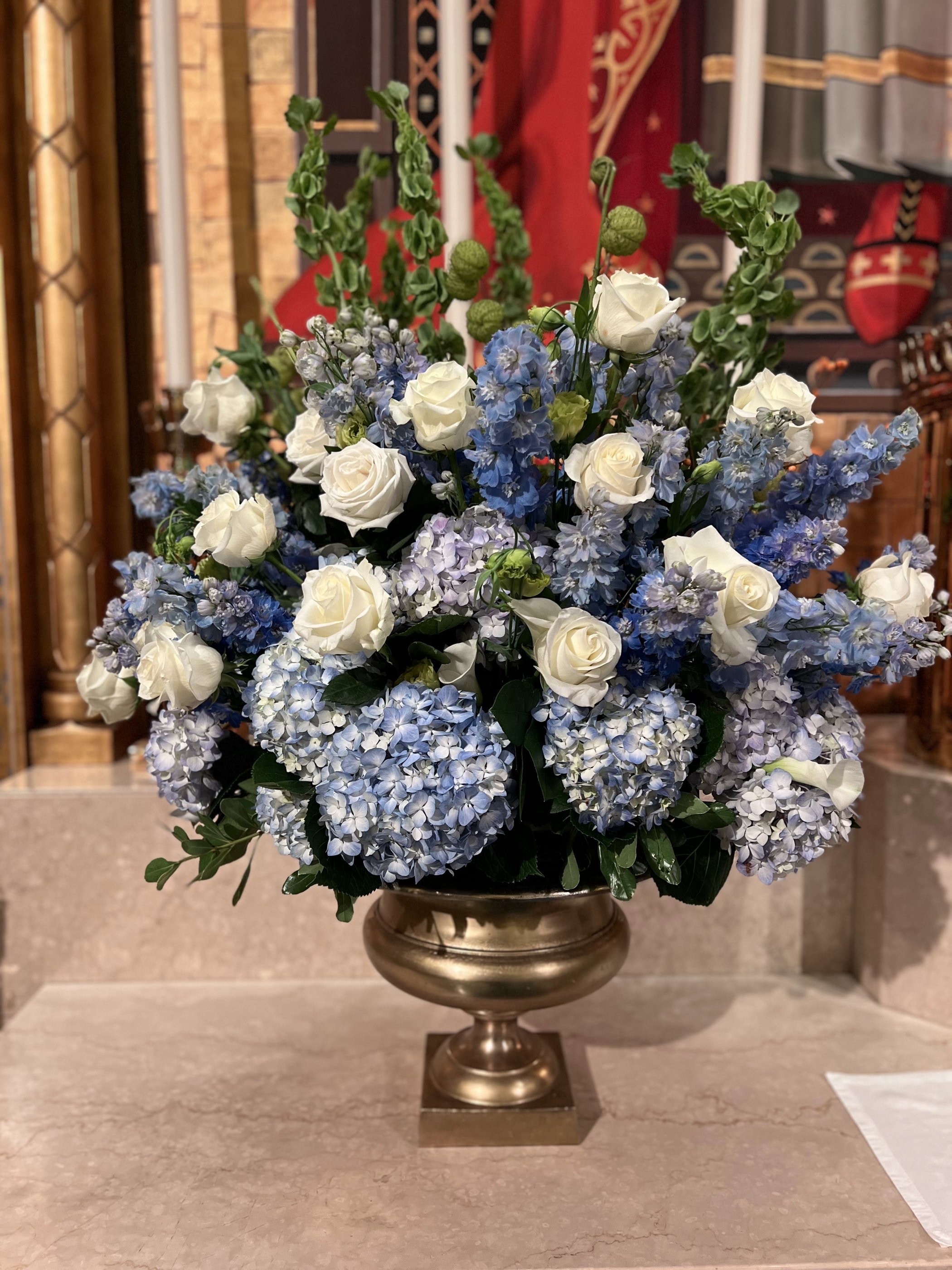 A blue and white arrangement of hydrangea, delphinium and roses in a brass urn