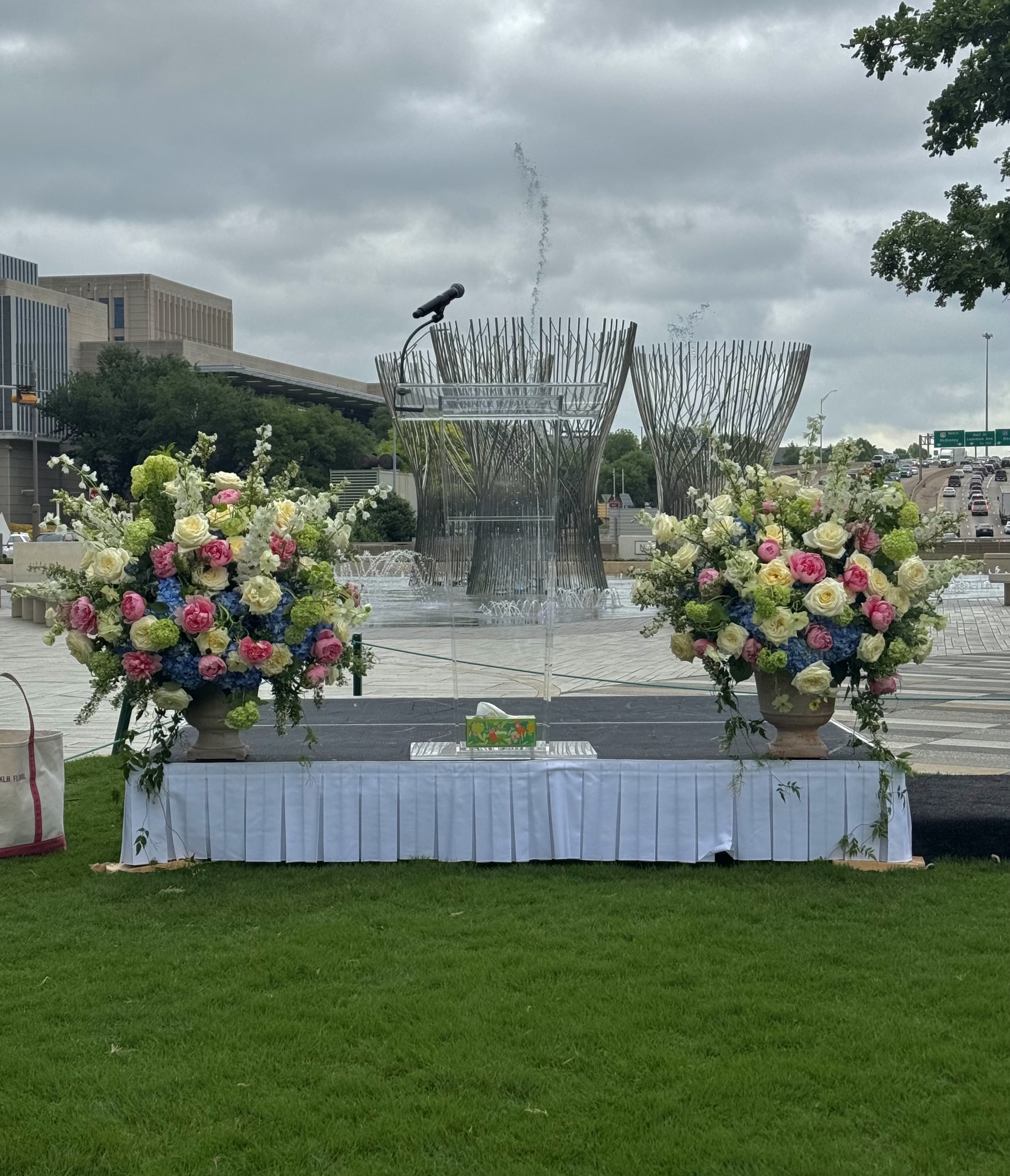 Two tall urn arrangements of pink and white blooms flanking an outdoor lectern beside a park fountain