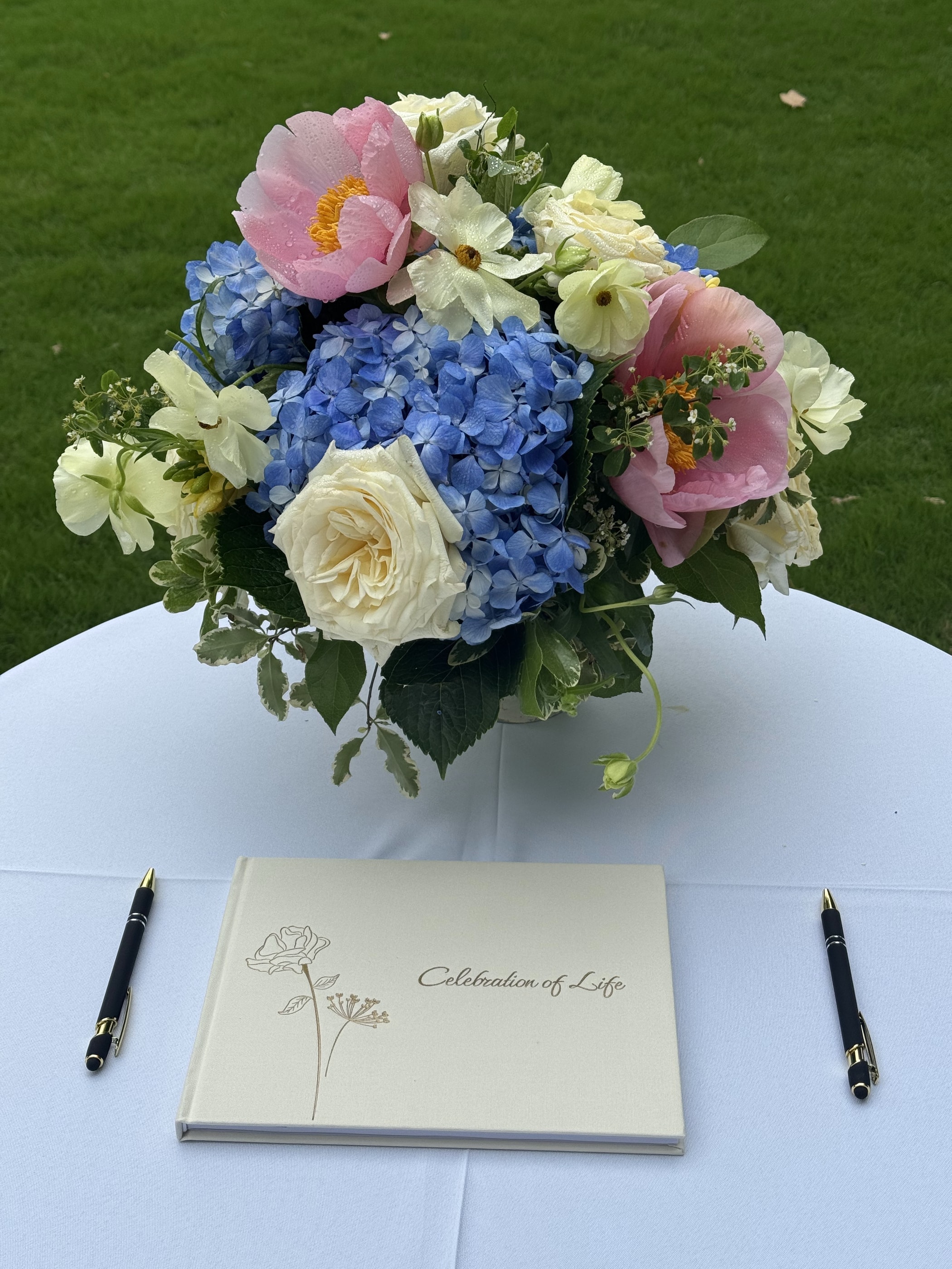 A pink and blue arrangement of peonies, hydrangea and narcissus beside a Celebration of Life guest book