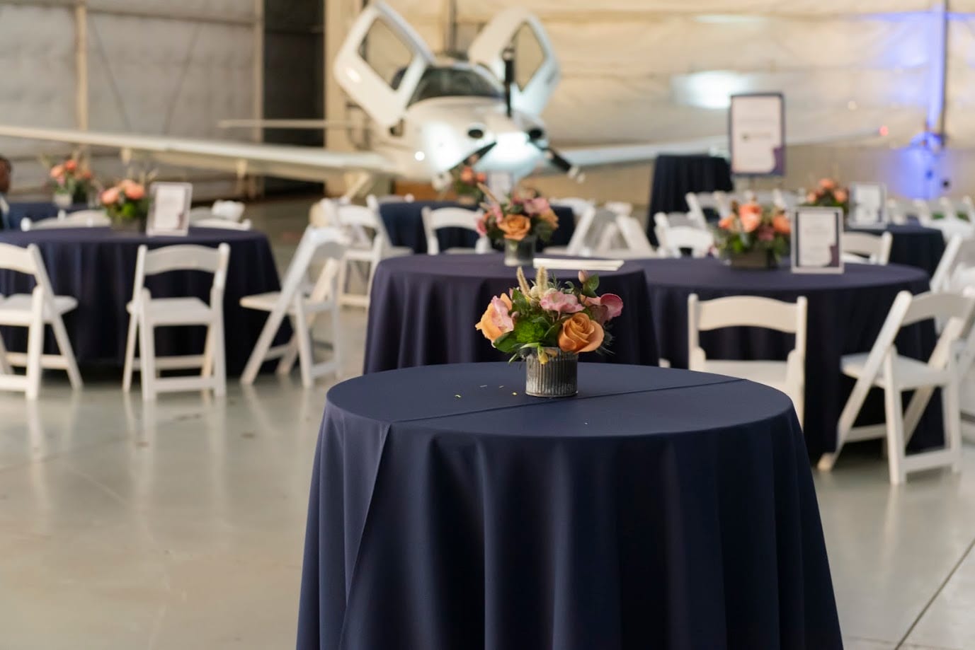 Fundraiser cocktail tables in navy linens with peach and coral arrangements inside an airplane hangar