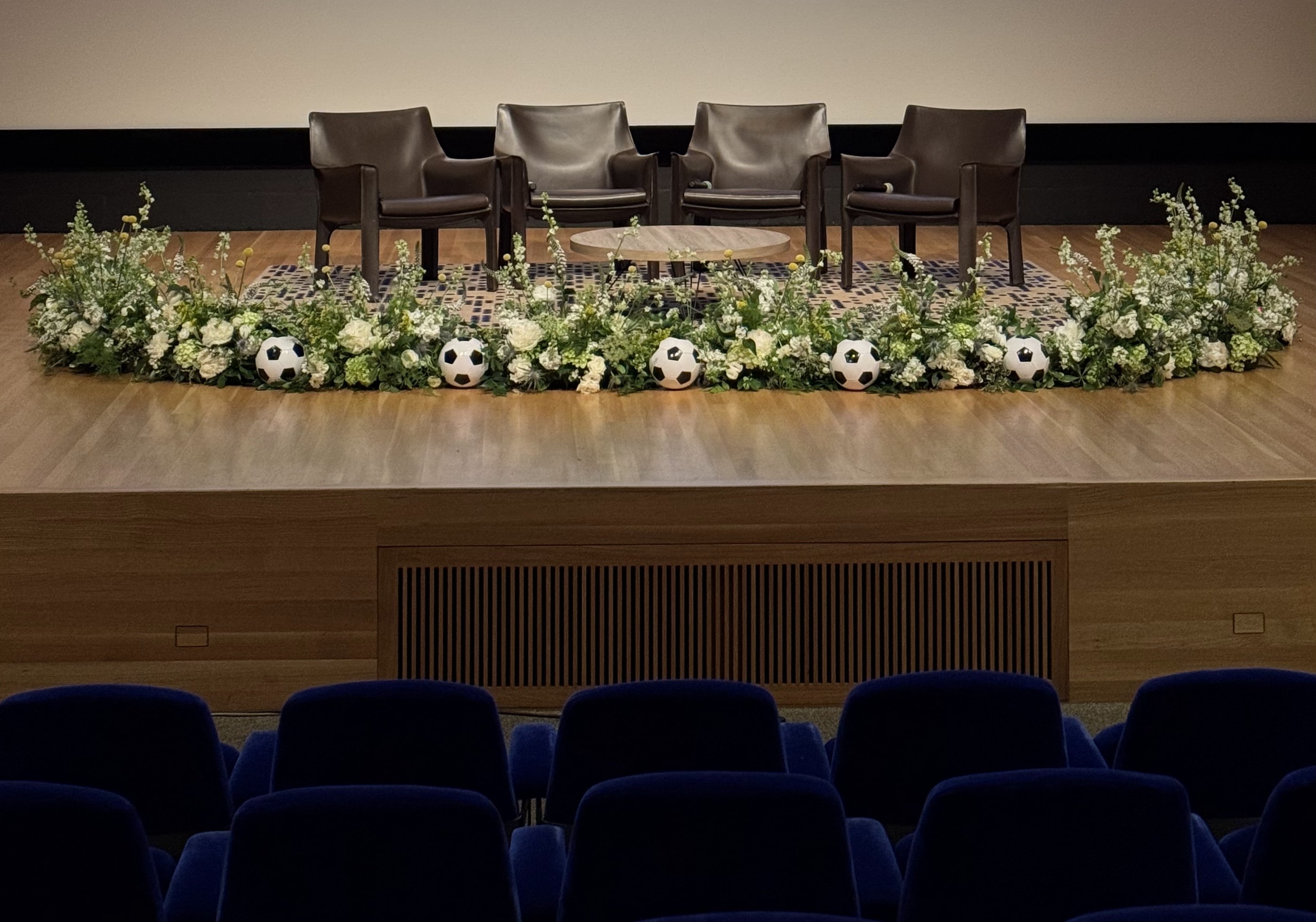 A long white and green garland running the front of an auditorium stage, punctuated with soccer balls beneath four panel chairs