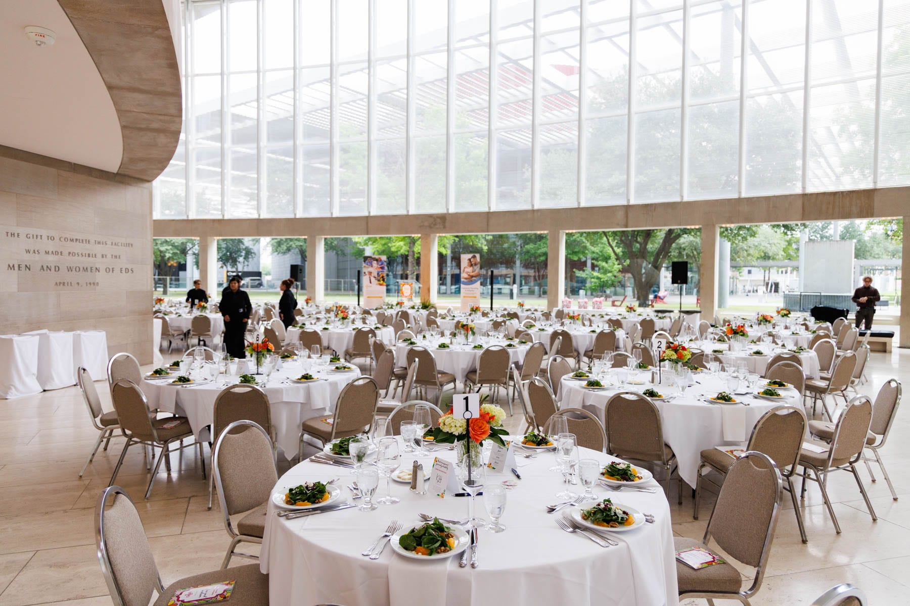 Luncheon tables under a bright clerestory atrium, each set with a low orange and peach centerpiece