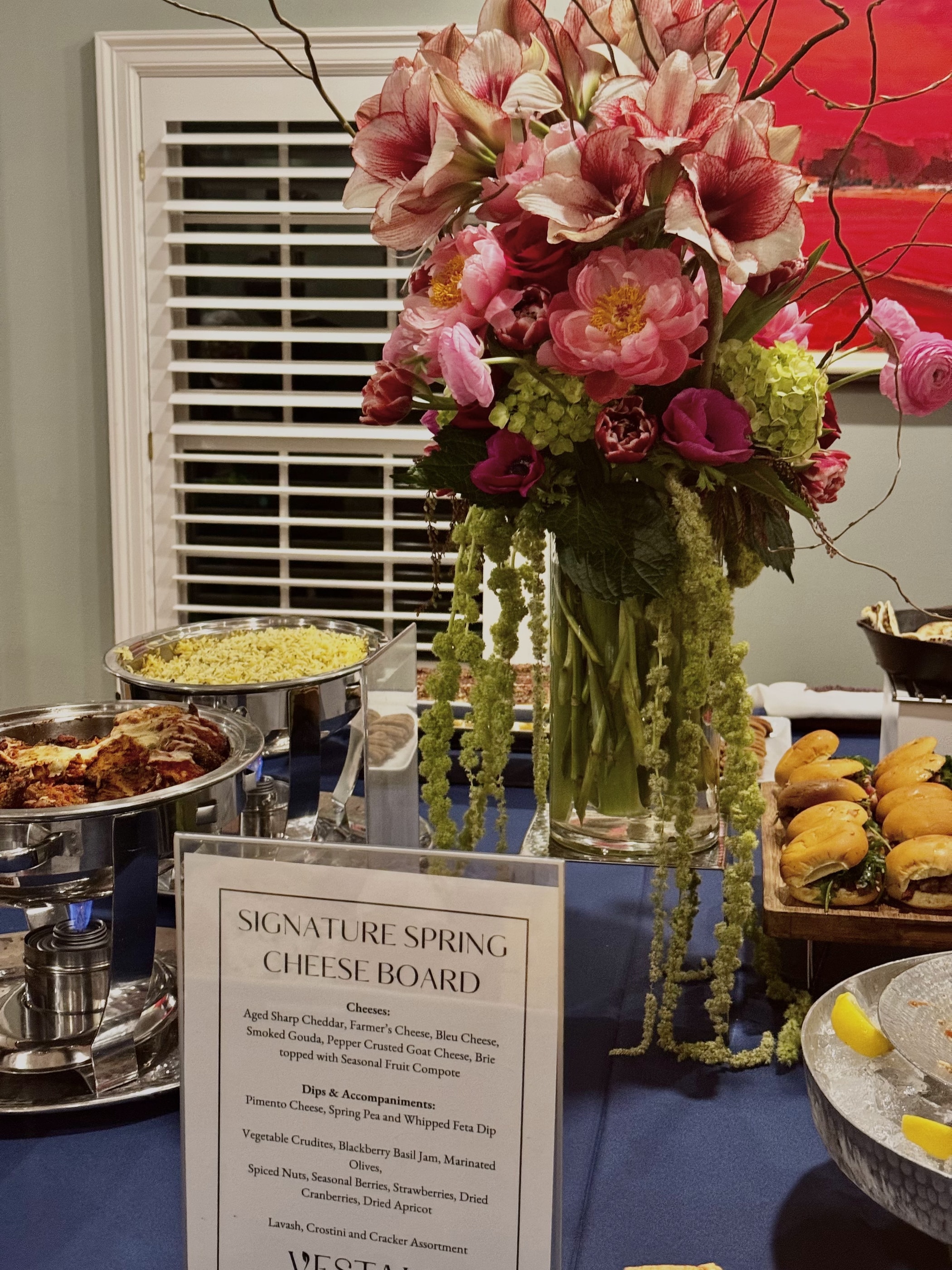 A tall buffet arrangement of pink amaryllis, peonies and green hydrangea with trailing amaranthus beside a cheese board