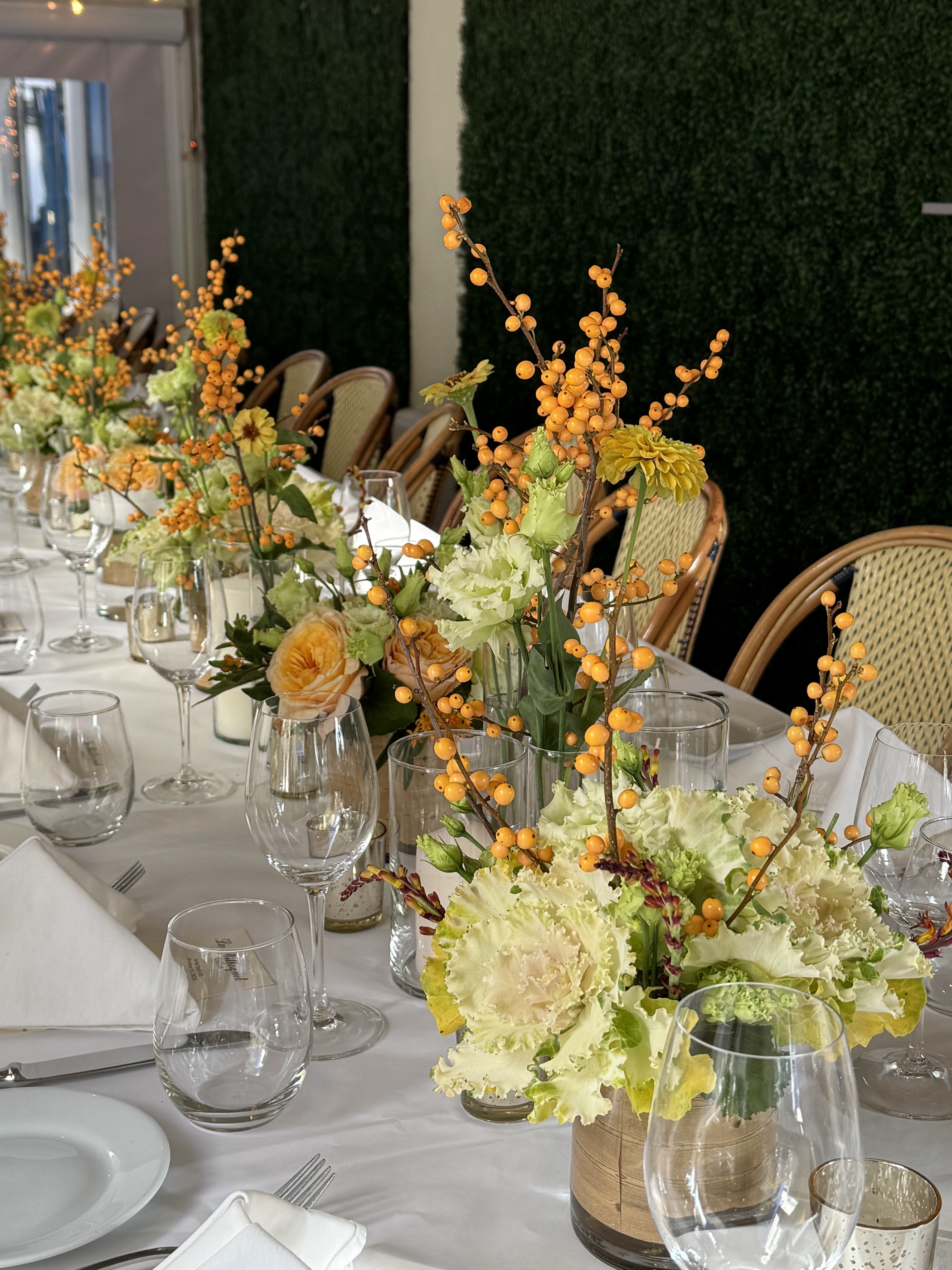 A long table against a boxwood hedge set with orange bittersweet branches, ornamental kale and golden roses
