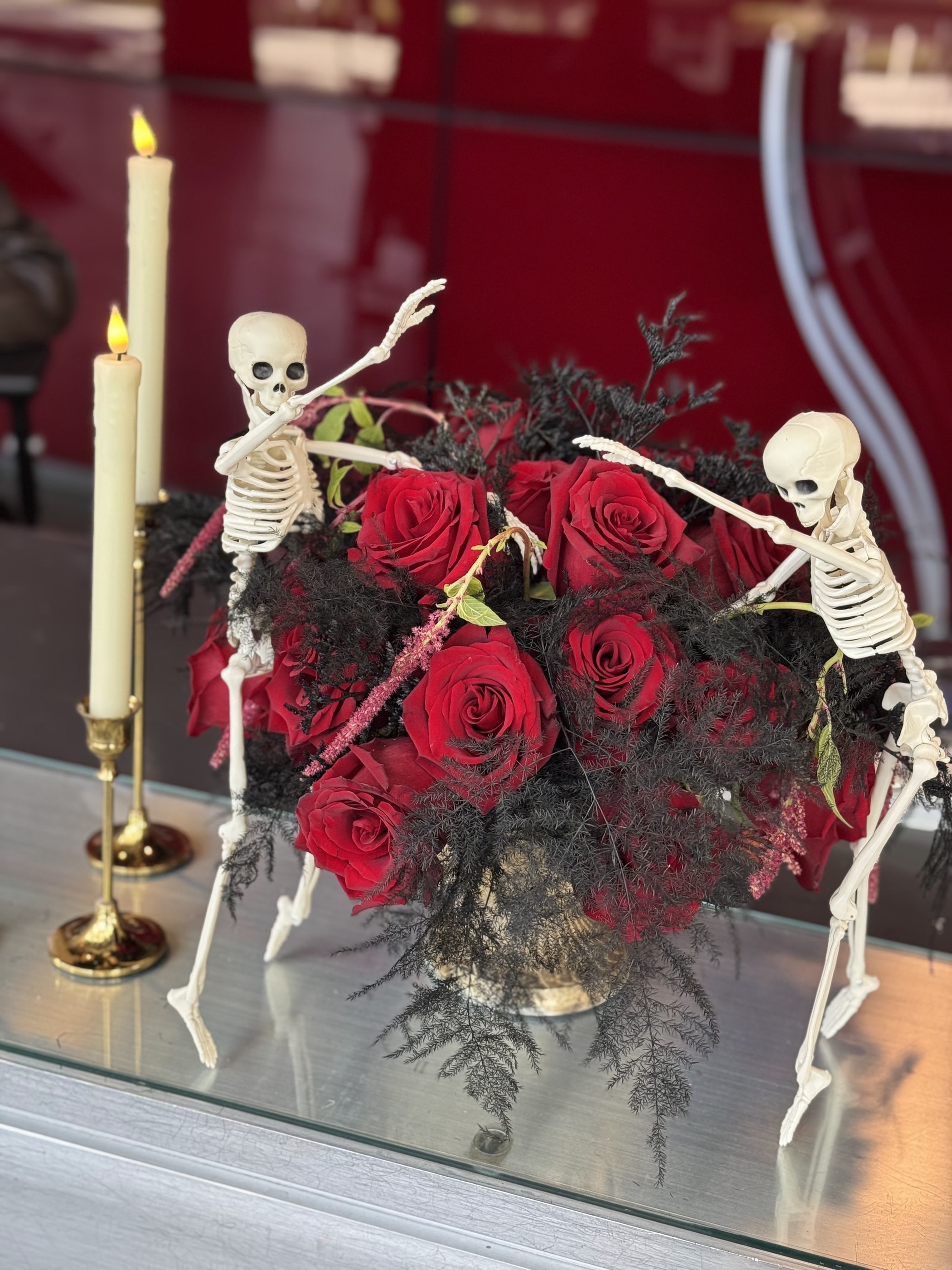 A Halloween arrangement of deep red roses and black foliage in a gold compote, flanked by taper candles and posable skeletons