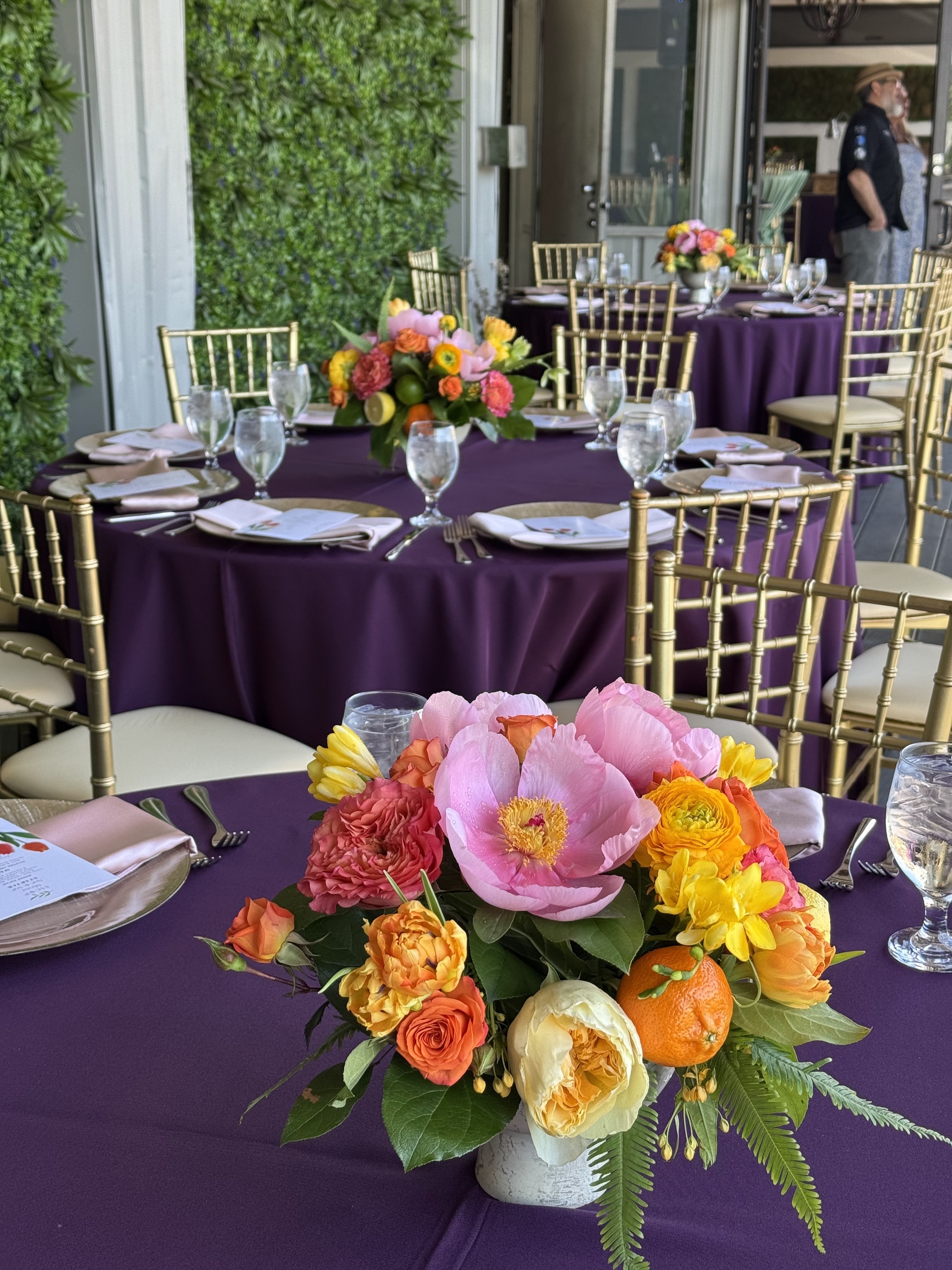 Luncheon tables in plum linens with gold chairs and bright pink peony, orange ranunculus and citrus centerpieces against a boxwood wall
