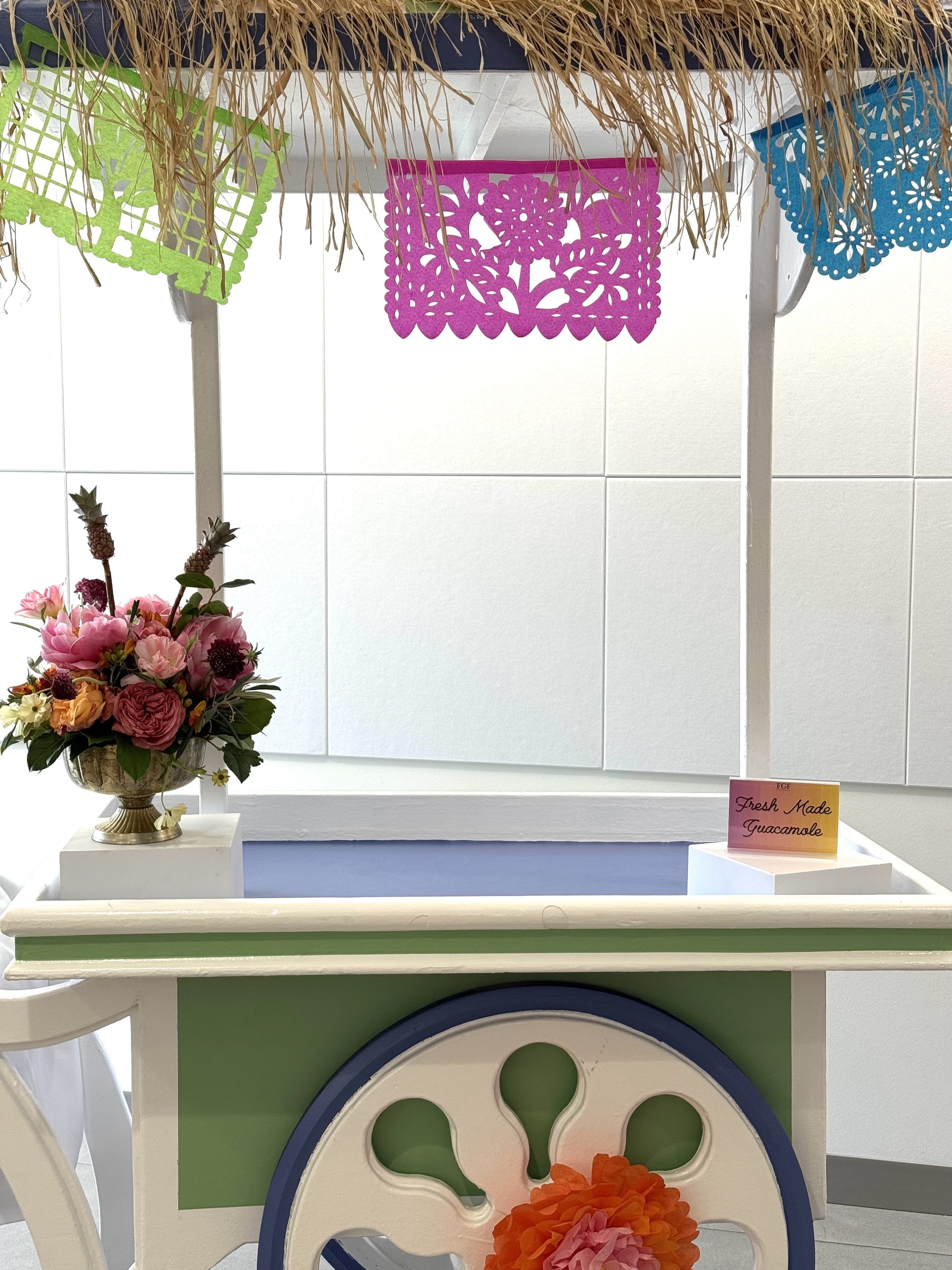A festive taco cart under papel picado banners with a pink and burgundy arrangement on the counter