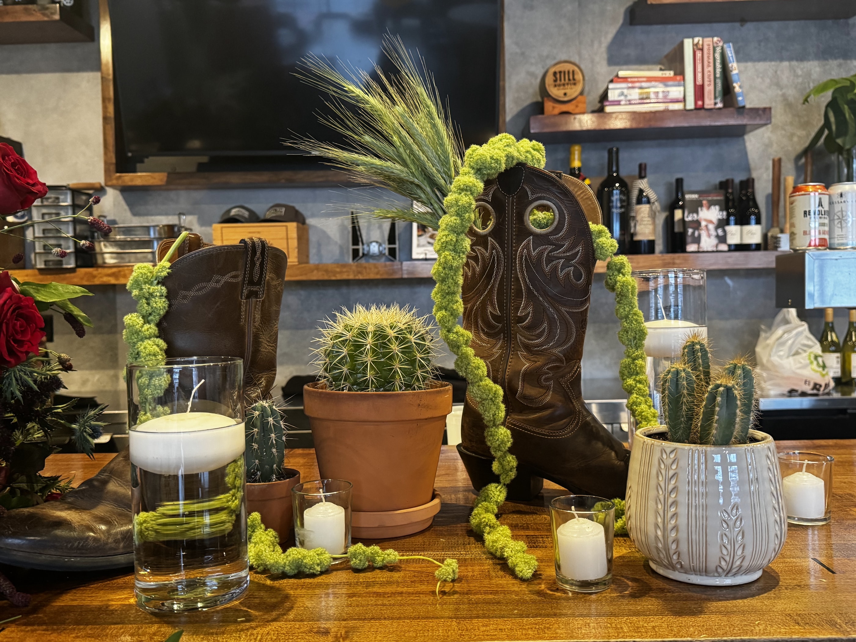 A western-themed bar top styled with cowboy boots, potted cacti, trailing green amaranthus and pillar candles