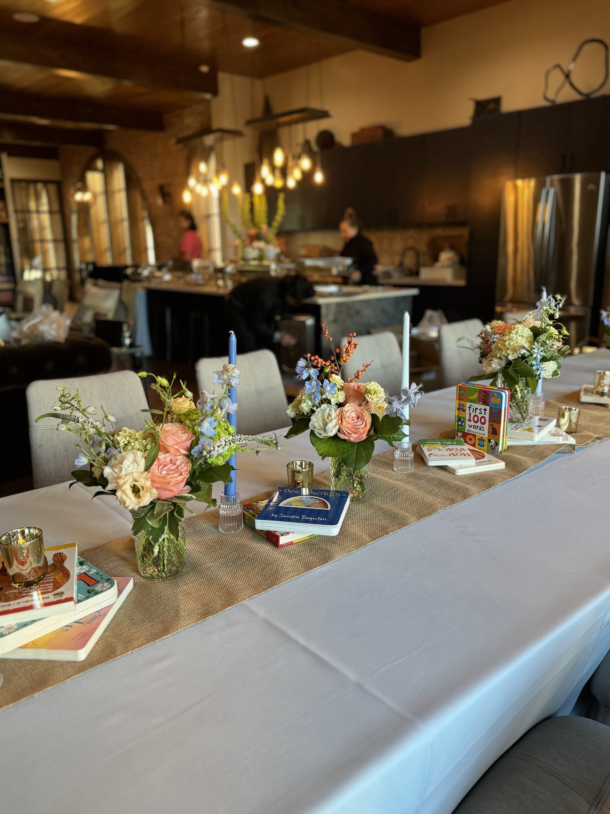 A baby shower table with a burlap runner, blue taper candles, small peach and blue bud arrangements and stacked children's books