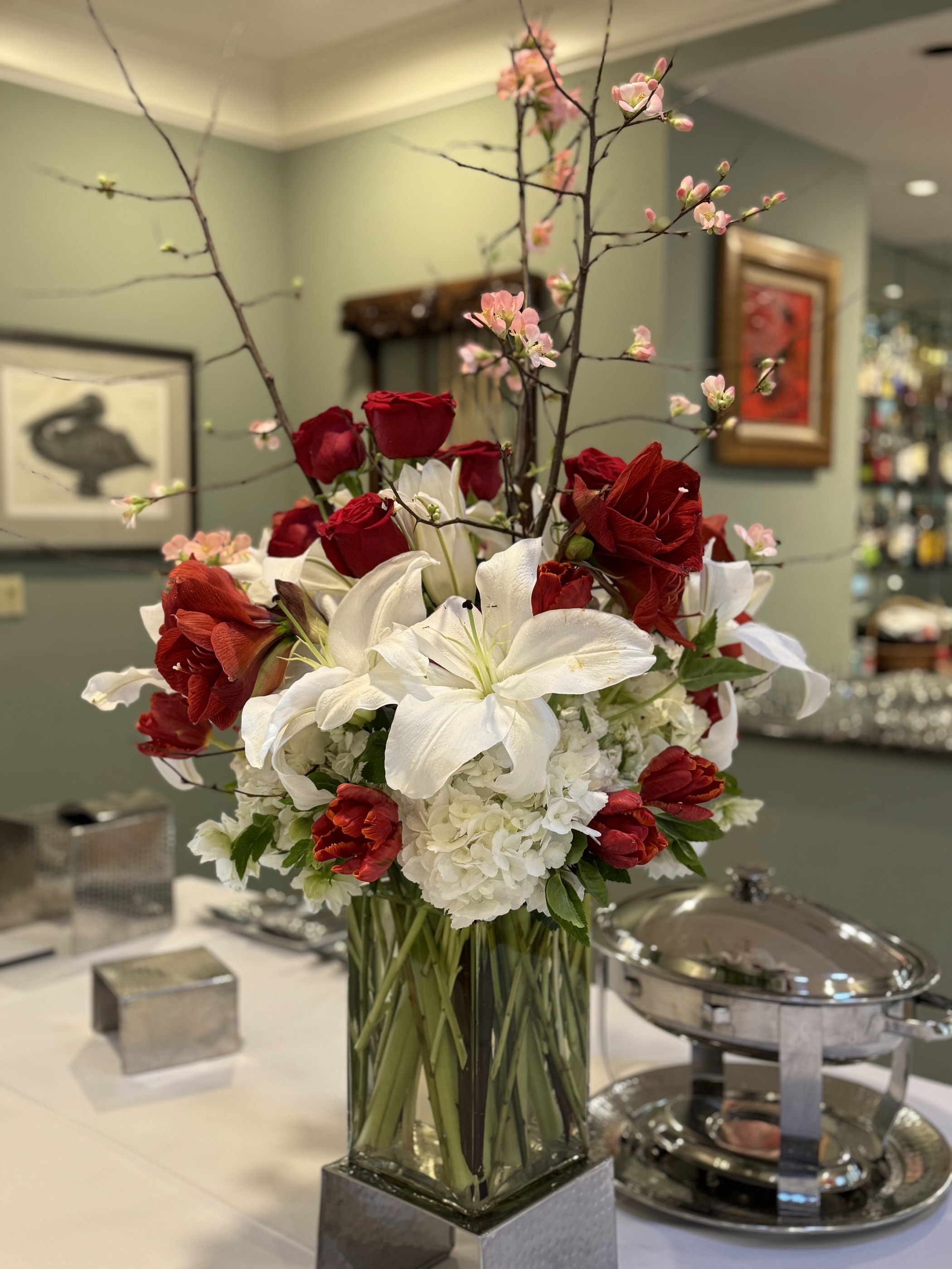 A tall holiday arrangement of red roses, amaryllis, white lilies and blossoming branches on a buffet