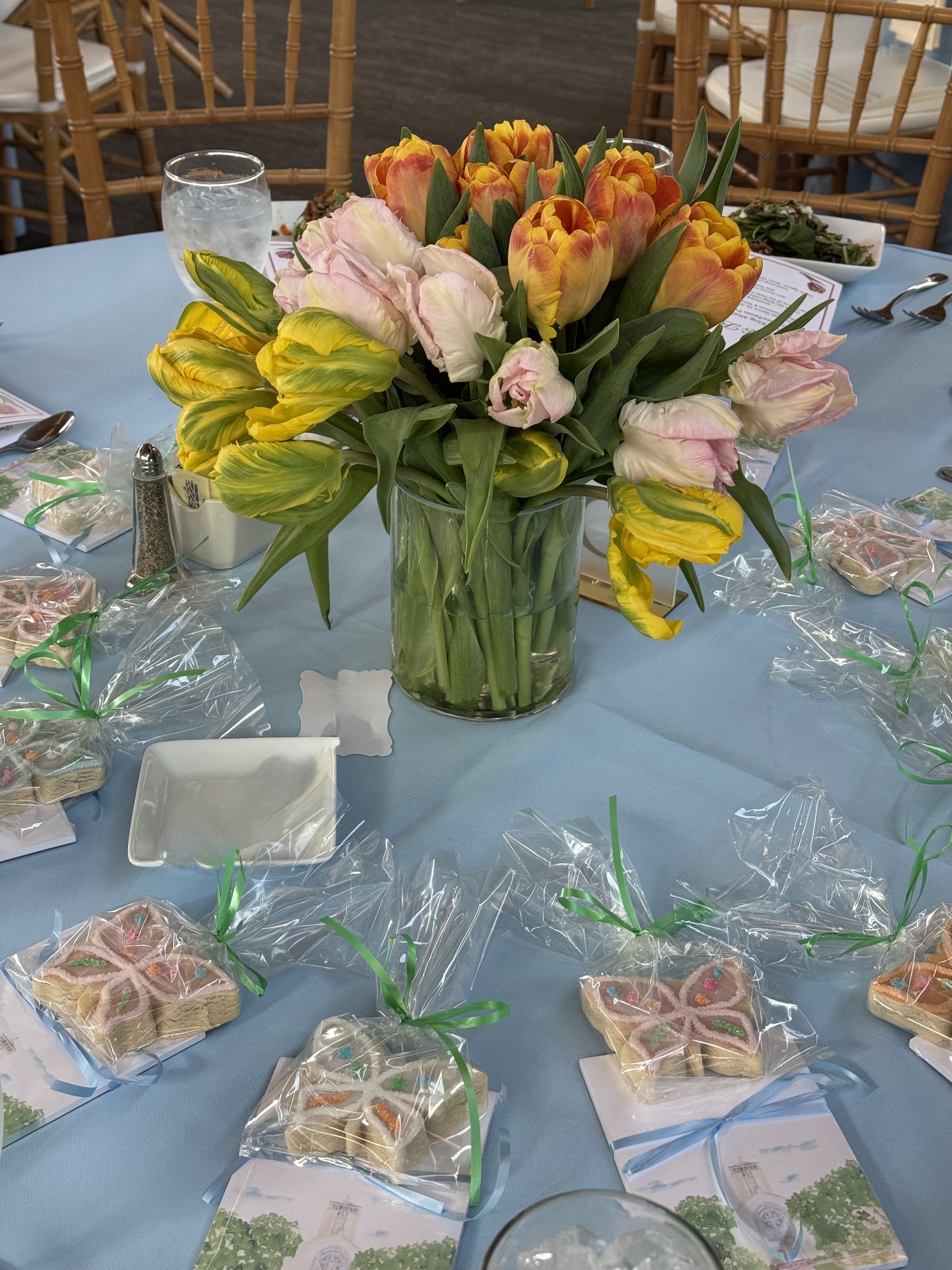 A clear vase of orange, yellow and pink parrot tulips on a pale blue table surrounded by ribbon-tied decorated cookies