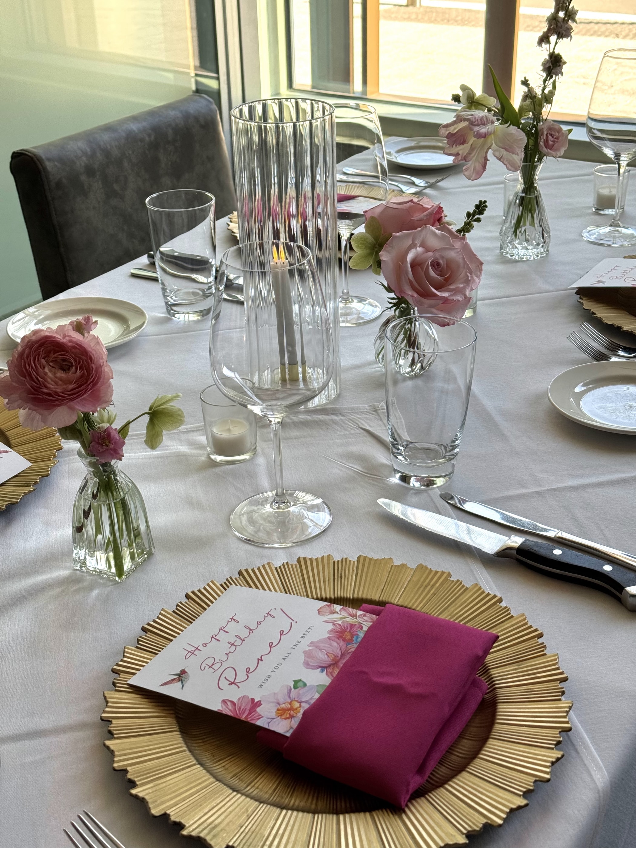 A birthday place setting with a gold charger, magenta napkin and a hand-lettered card, with pink rose and ranunculus bud vases along the table