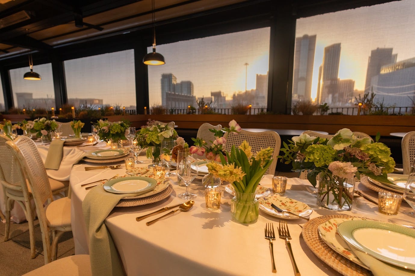 A long celebration table at golden hour with the Dallas skyline behind it, low green and white arrangements and yellow tulips down the center