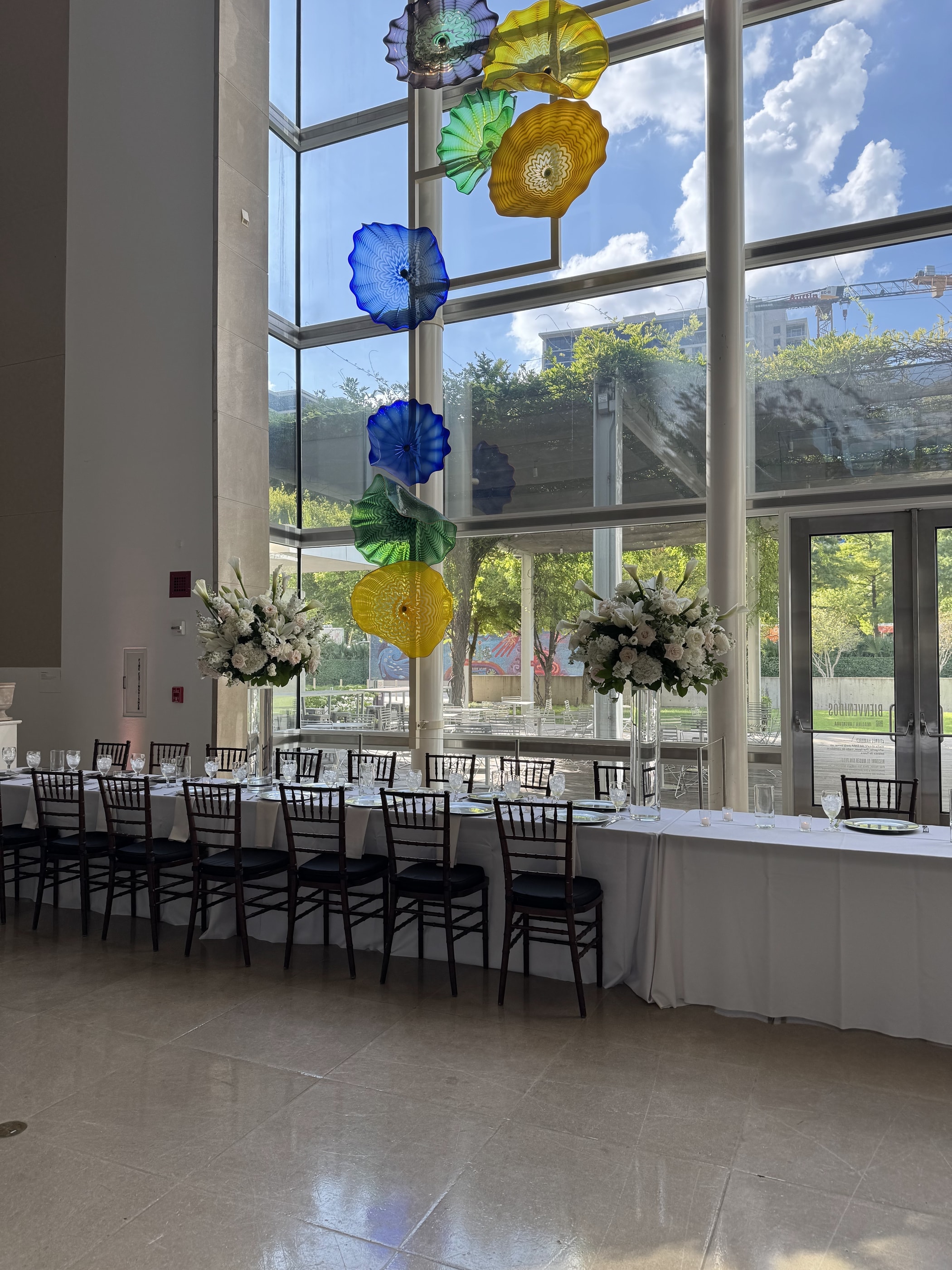 Tall white and green arrangements beside a long dinner table at the Dallas Museum of Art, with colorful blown-glass sculpture in the window