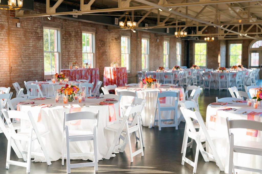 Reception in a brick-walled venue with round tables, white folding chairs and bright coral and orange centerpieces