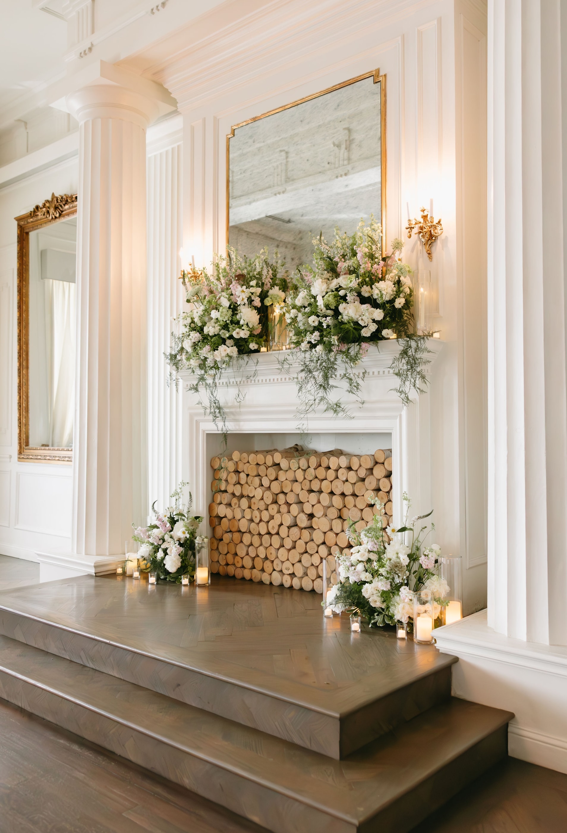 A white marble mantel framed with a lush white and blush floral installation, trailing greenery and pillar candles at The Mason