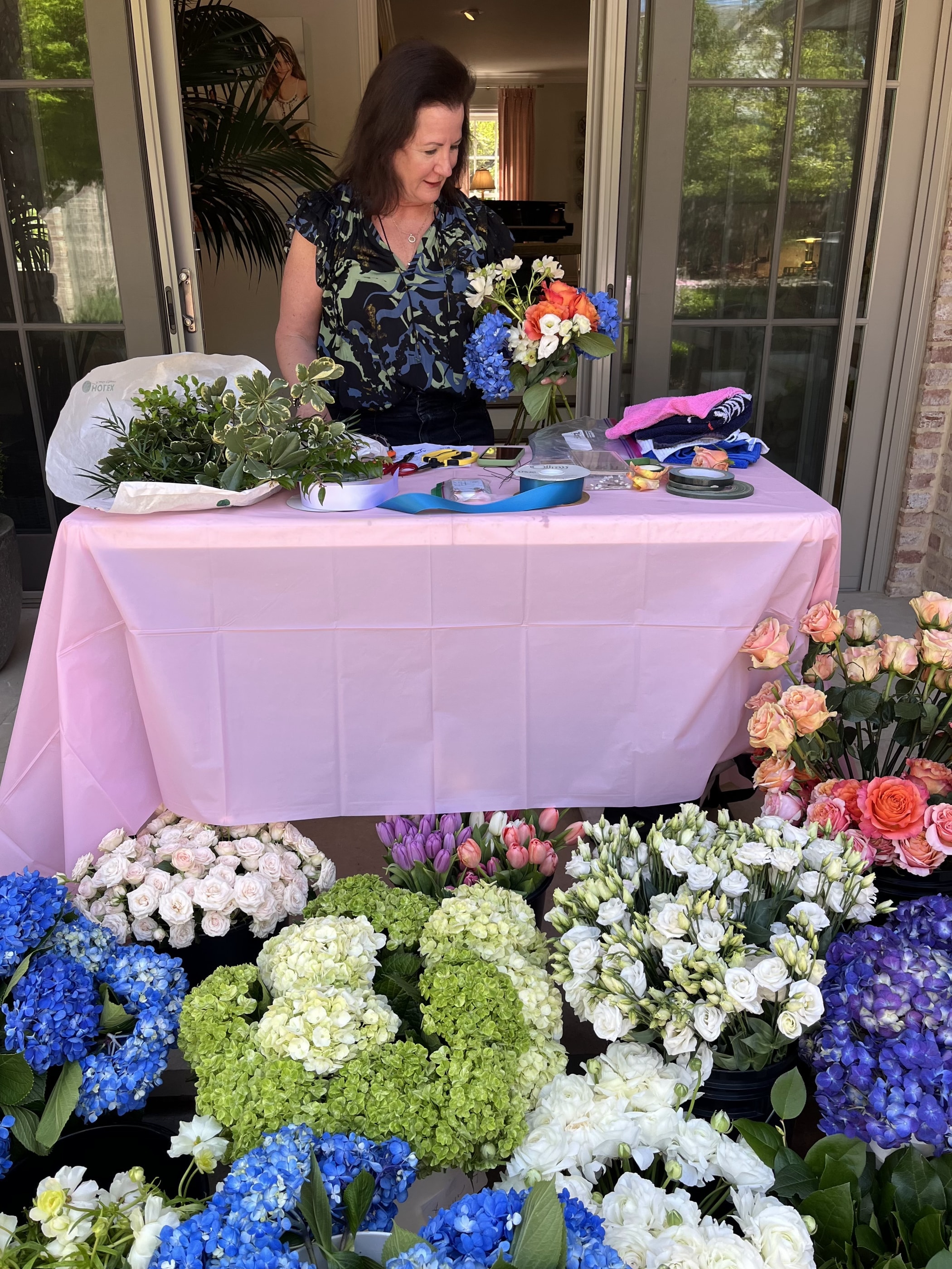 A florist building an arrangement at a draped worktable, with buckets of blue hydrangea and roses in the foreground