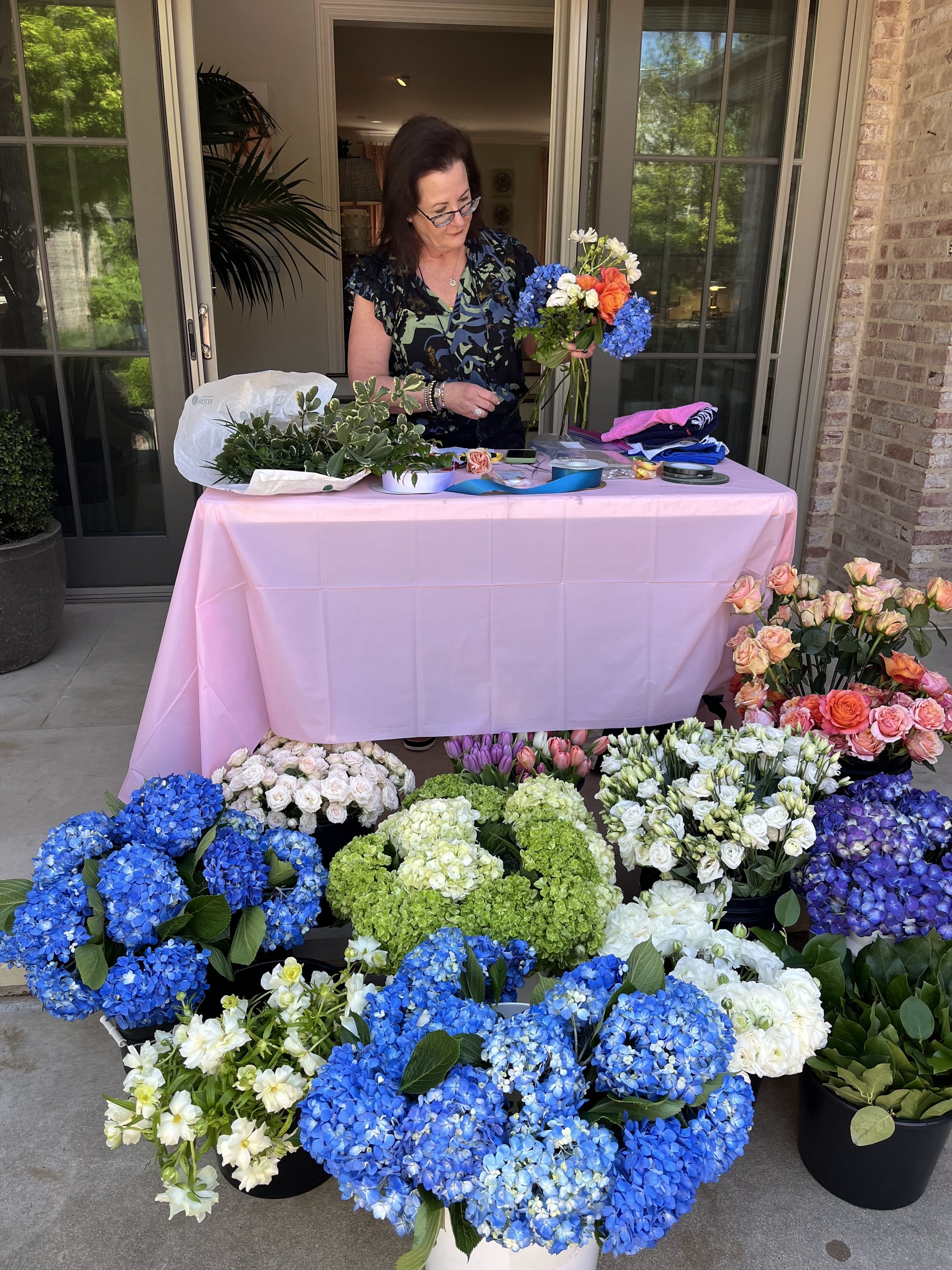 Buckets of blue and white hydrangea, roses and tulips banked in front of a workshop table as an arrangement is built
